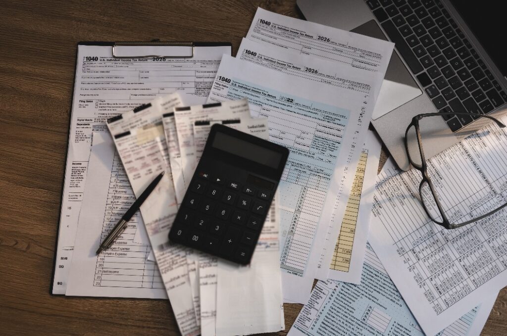 Steuerformulare, Belege und ein Taschenrechner, angeordnet auf einem Schreibtisch zusammen mit Finanzunterlagen und einem Laptop, die die Steuererklärung, die Buchhaltungsprüfung, die Ausgabenberechnung und den Arbeitsablauf bei der Steuererklärung unterstützen.